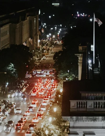 A nighttime cityscape showing a busy street lined with cars and illuminated buildings, with flags visible and soft street lighting.